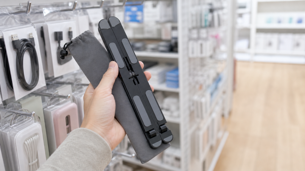 100円ショップのPC周辺機器売り場で黒い折りたたみ式パソコンスタンドを探すイメージ